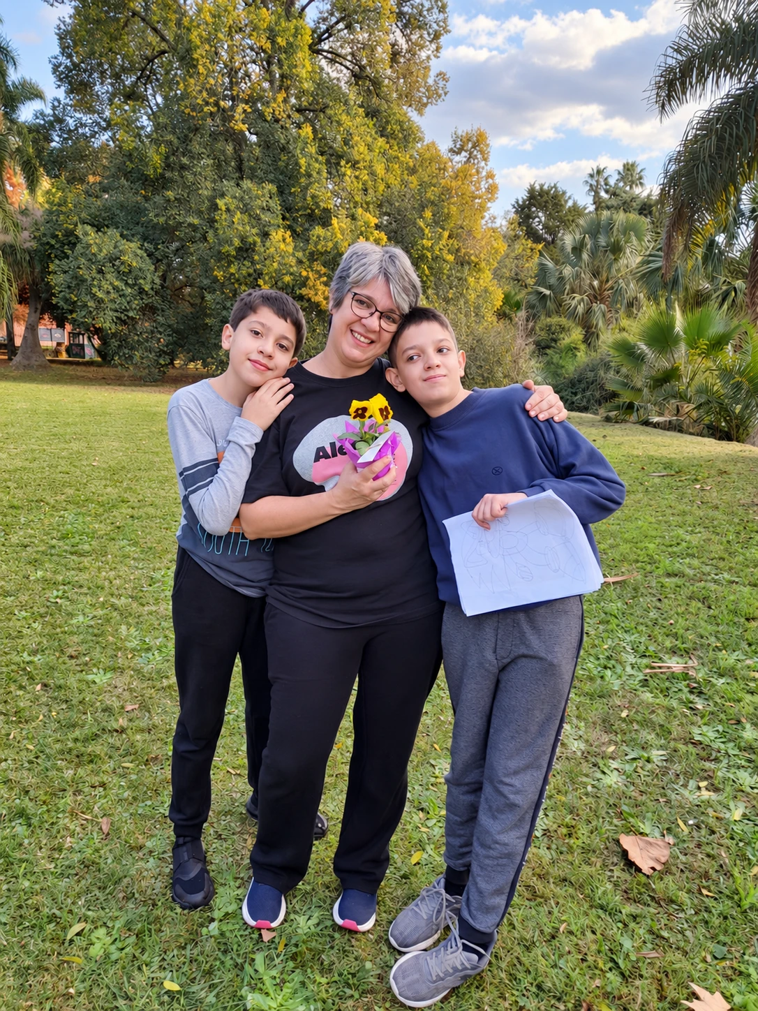 Tres personas posan en un jardín; una sostiene flores y otra un dibujo.