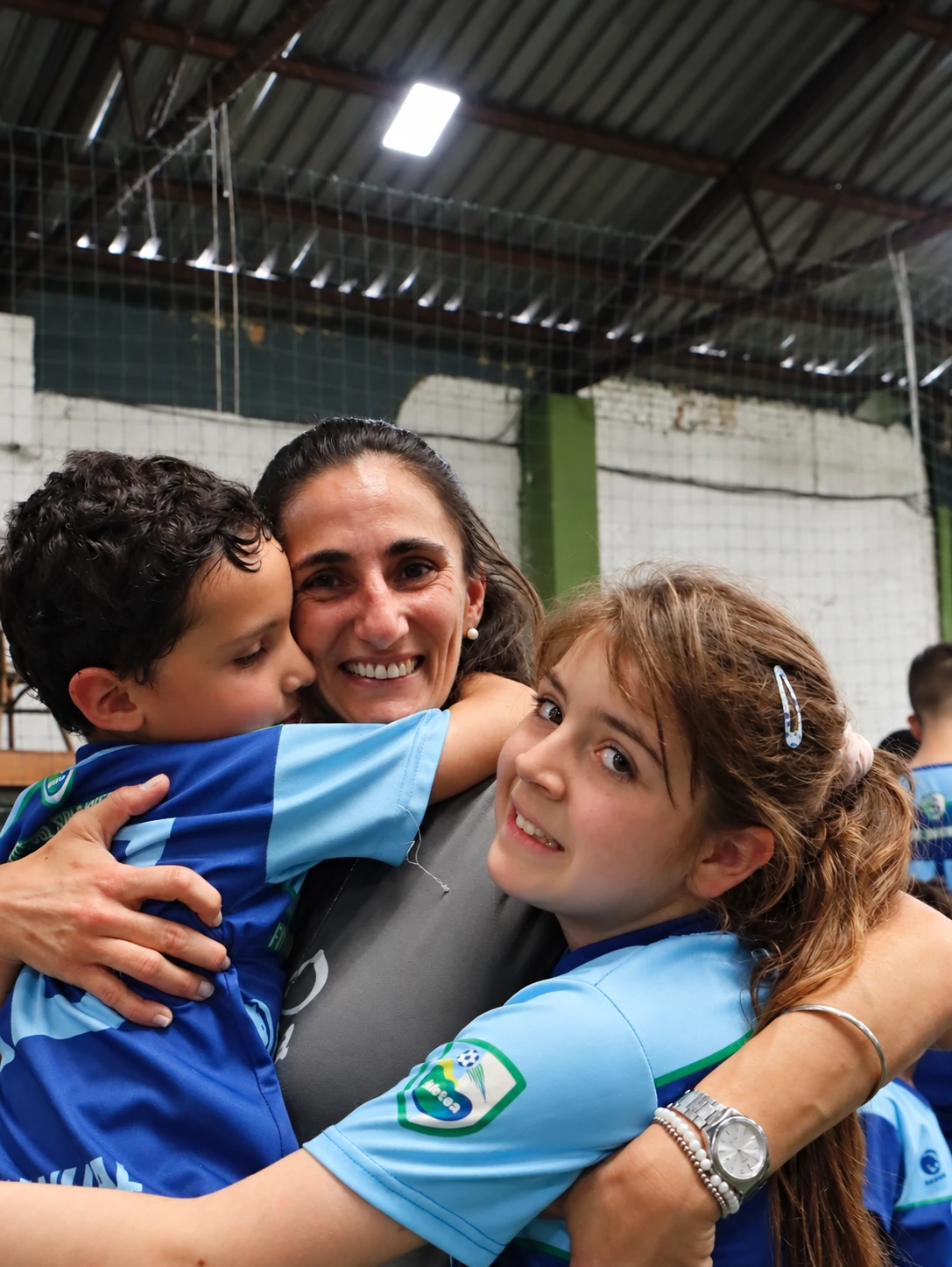 Una persona adulta abrazada por dos niños con camisetas azules en un gimnasio.