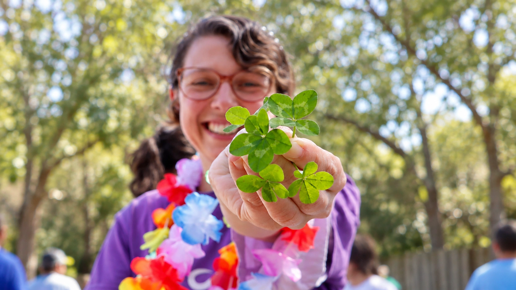 Persona sonriente con gafas y camiseta violeta ofrece tréboles con la mano, al aire libre y con collares de flores.